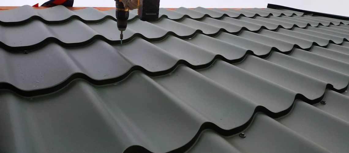 The professional worker works on installation of a roof of a roof by sheets of a metal tile and drills a screw with a drill
